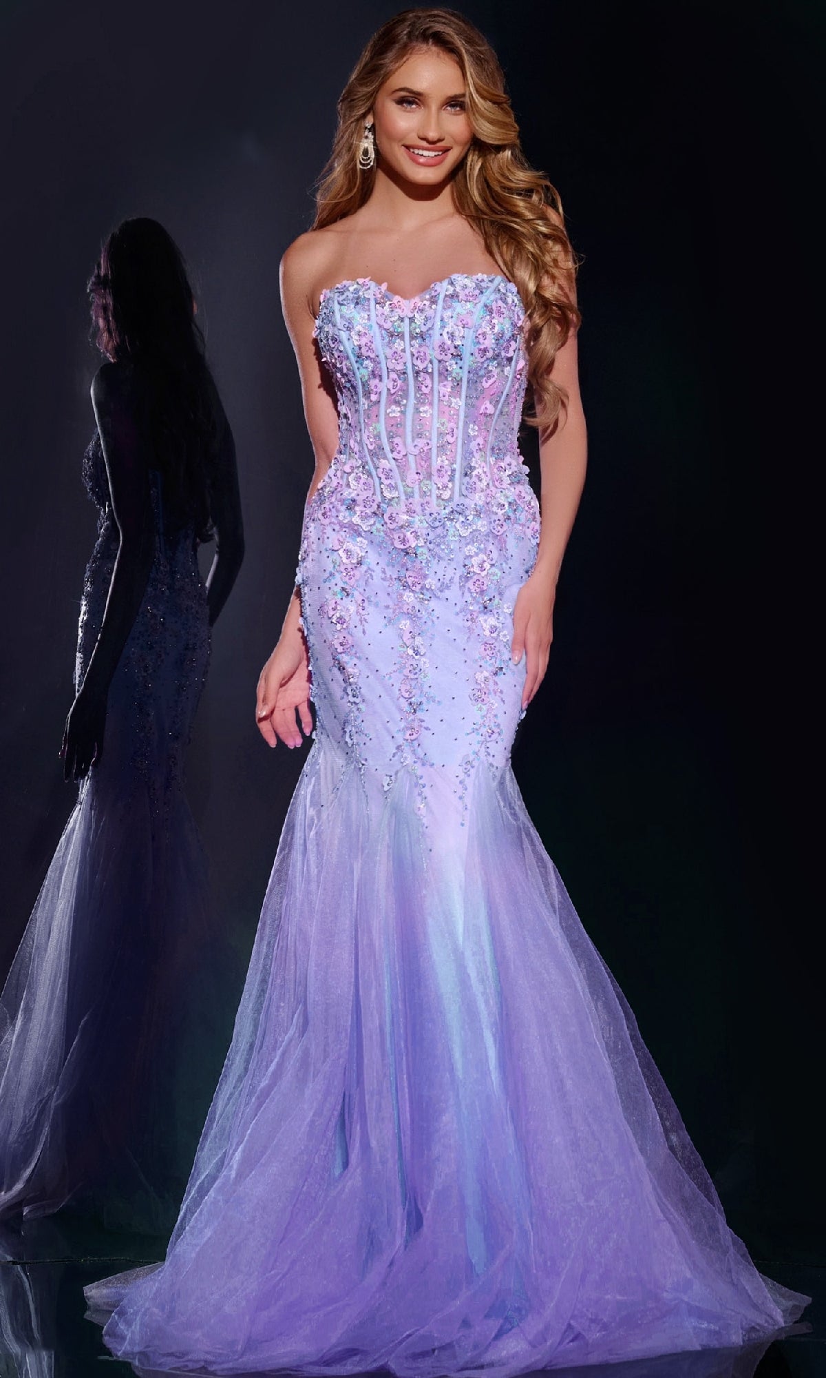 A woman wearing the Strapless Long Mermaid Prom Dress Jovani 44211 in lavender with floral embellishments stands smiling, her reflection visible behind her.